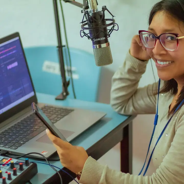 Podcaster in headphones smiling at a boom microphone, audio software open on her laptop