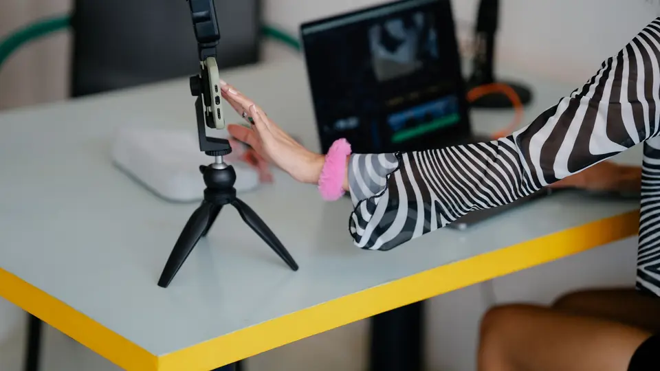Creator at a yellow desk reaching for her phone on a tripod, video edit open on the laptop